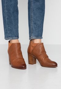 Bottines marron avec une texture tissée sur le devant, des accents en daim lisse et un talon empilé, associées à un jean bleu coupé.