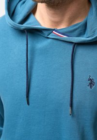Tyrkysová bavlněná mikina s kapucí, stahovací šňůrkou a vyšívaným logem. Má kapuci a texturovanou látku s kontrastními detaily u krku.