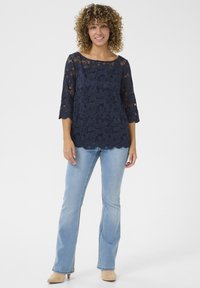 Top in pizzo navy con maniche a tre quarti, motivo floreale, scollo rotondo e orlo scallopato, abbinato a jeans a zampa d'elefante blu chiaro.