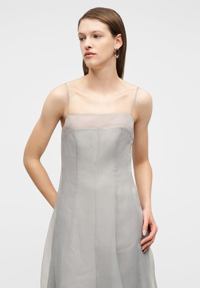 Νεαρή γυναίκα με ίσια καστανά μαλλιά φοράει ανοιχτό γκρι αμάνικο φόρεμα με λεπτούς ιμάντες και χρυσούς κρίκους σκουλαρίκια σε φόντο απλό λευκό.