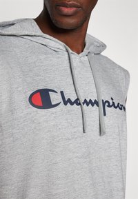Grijze mouwloze hoodie met een groot "Champion"-logo in marineblauw met een rode accent, gemaakt van een zachte, gestructureerde stof met een trekkoord.