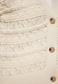 Cardigan in crema lavorato a maglia con motivi testurizzati, bordi a coste e grandi bottoni marroni. Il motivo include diamanti e cuori.