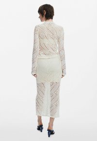 Vit spetsklänning med långa ärmar och en figurnära silhuett, med ett geometriskt mönster och knälång fåll. Stylist med blå klacksandaler.
