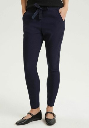 Osoba nosząca dopasowane granatowe spodnie dresowe z regulowanym paskiem w talii i czarne baleriny typu Mary Jane, stojąca z rękami w kieszeniach.