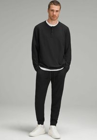 Zwarte langemouwen henley shirt met drie knopen, gecombineerd met zwarte joggerbroeken en witte sneakers. Glad materiaal, relaxte pasvorm, minimalistisch ontwerp.