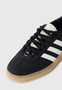 Black athletic shoe with white stripes, featuring a textured rubber sole and black laces. The upper is made of smooth material. Gold logo detail.