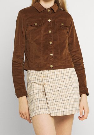 Kvinde iført en brun kordfløjlsjakke med knapper og en beige ternet mininederdel med knapper foran, stående mod en ensfarvet baggrund.