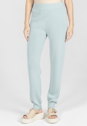 Jasnoniebieskie dzianinowe spodnie dresowe z prążkowanymi ściągaczami, noszone z beżowymi sandałami na platformie, na jednolitym tle.