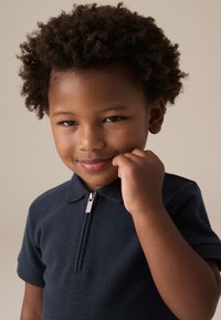 Polo bleu marine à manches courtes, col et détail en fermeture éclair argentée. Le tissu a une apparence texturée. La main d'un enfant est levée vers le visage.