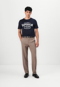 Marineblauwe katoenen t-shirt met witte bedrukte tekst, gecombineerd met lichtbruine broeken en zwarte schoenen, wat een casual, getailleerde stijl toont.