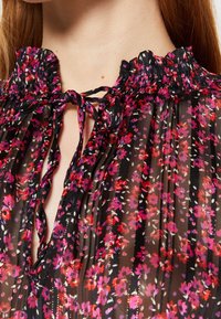 Blusa nera floreale con colletto arricciato, caratterizzata da piccoli fiori rosa e bianchi, chiusura con laccetto al collo e tessuto trasparente e texturizzato.
