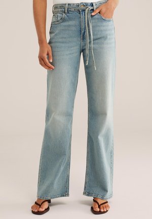 Osoba nosiaca svetlomodré široké rifle so svetlo bielym opaskom na zaväzovanie a tmavohnedými sandálmi na podpätku, stojaca na jednofarebnom pozadí.