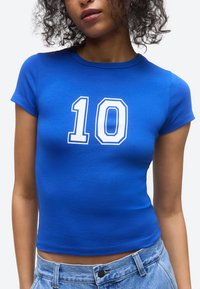 Naine, kes kannab vormikat sinist t-särki, millel on valge number "10" rinnal, kombineeritud helesiniste teksadega.