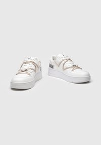 Zapatillas blancas bajas con cordones beige, suelas gruesas y texto negro "BULK MAGH" en el lateral, mostradas sobre un fondo gris liso.