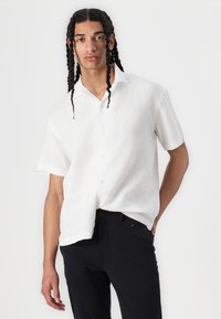 Chemise blanche en lin à manches courtes avec col et boutons, portée avec un pantalon noir. La chemise présente une coupe décontractée et un tissu texturé.
