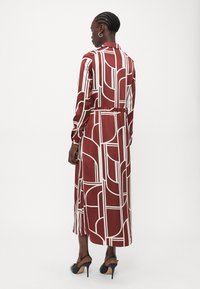 Lungo vestito color bordeaux con motivi geometrici bianchi, maniche lunghe e vita con cintura. Tessuto simile alla seta con taglio dritto e design alla caviglia.