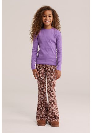 Jeune fille aux longs cheveux bouclés portant un t-shirt violet à manches longues, un pantalon évasé marron à motifs et des chaussures marron, souriant à la caméra.