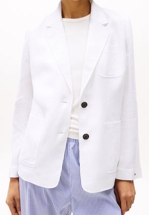 Persoon draagt een witte blazer met twee zwarte knopen over een witte top en blauw-wit gestreepte broek.