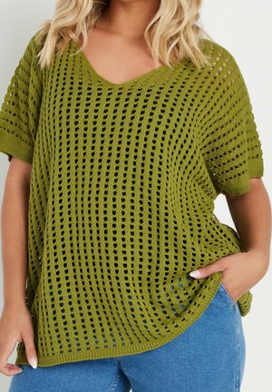 Femme portant un haut tricoté vert olive à manches courtes et amples, avec un motif ajouré, sur un jean bleu, main dans la poche.