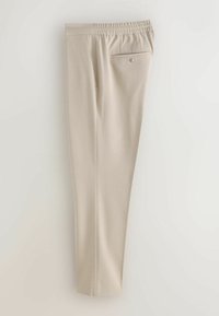 Pantalon beige fuselé avec ceinture élastique et une poche arrière unique sécurisée par un bouton, présenté sur un fond uni.