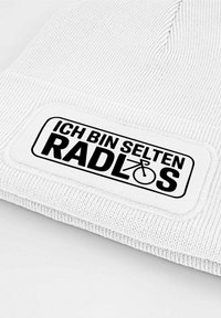 White knit beanie with a textured finish. Features a black rectangular patch with the text "ICH BIN SELTEN RADLOS" and a bicycle icon.