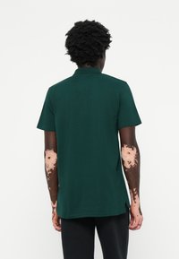 Camisa polo verde de mangas curtas e colarinho. O tecido parece texturizado. As costas apresentam fendas laterais para facilitar o movimento.