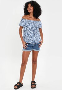 Blå off-the-shoulder bluse med blomsterprint og flæser ved halsen, parret med denimshorts med rullede manchetter og sorte slide-sandaler.