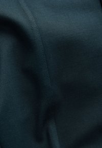 Nahaufnahme eines dunkel-türkisfarbenen Stoffes mit sichtbarer vertikaler Naht und subtiler Textur, die leichte Falten und ein glattes Gewebe zeigt.