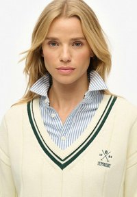 Cremfarbener Strickpullover mit V-Ausschnitt, versehen mit grünen Streifen, kombiniert mit einem blau-weiß gestreiften Kragen. Logo sichtbar.