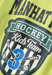 Das grüne Shirt verfügt über einen grafischen Druck mit dem Text "MANHATTAN" und "HOCKEY Kids Team", beinhaltet die Nummer "3" in Blau und gekreuzte Hockeyschläger.