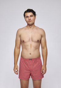 Rote karierten Boxershorts aus leichtem Stoff, mit einem elastischen Bund und einem Markenetikett auf der Vorderseite. Das Modell ist oberkörperfrei.
