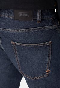 Donkerblauwe denim jeans hebben een achterzak met contrasterende oranje stiksels en een zwart label met de opdruk "camel active".