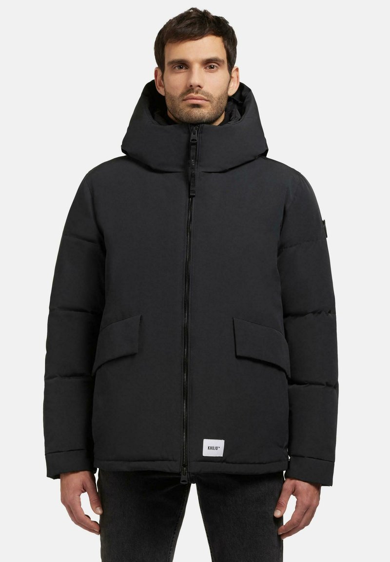 Schwarze gepolsterte Jacke mit hohem Kragen, Reißverschluss, zwei Vordertaschen und einem Etikett am unteren Saum. Glatte Textur, modernes Design.