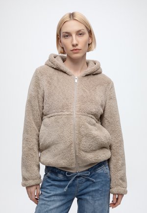 Femme aux cheveux blonds courts portant un sweat à capuche beige en peluche avec fermeture éclair et un jean bleu, debout devant un fond blanc uni.
