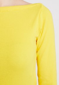 Gul räfflad långärmad topp med avskuren halsringning. Mjuk textur och figurnära passform, framhävd av framträdande räffling längs kanterna.