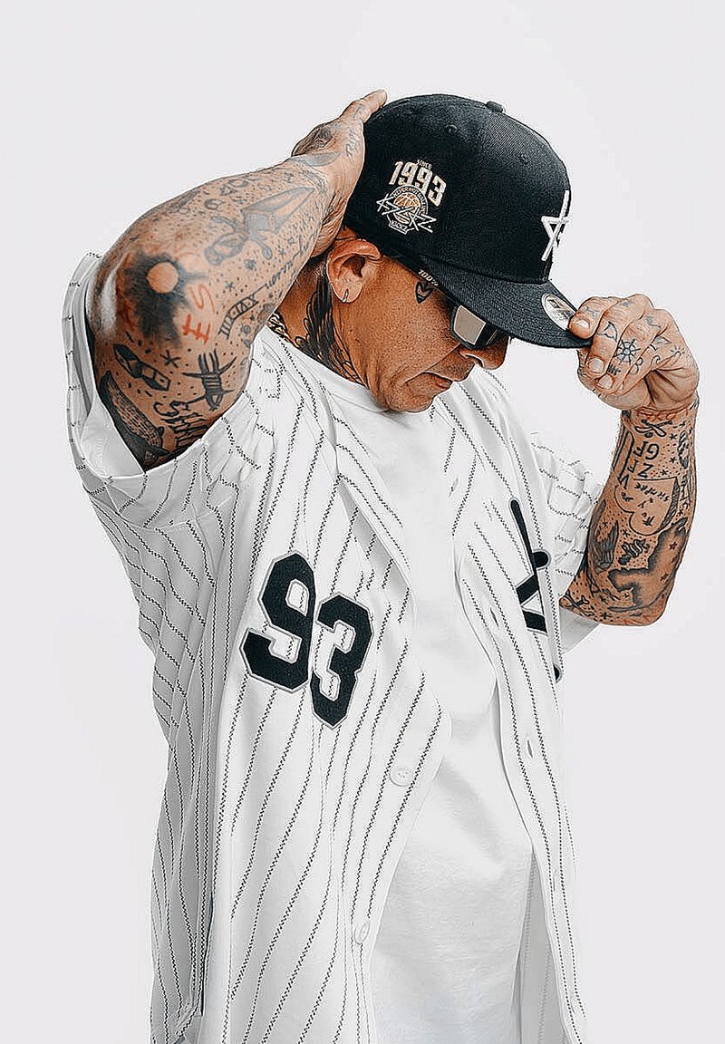Maglia da baseball con strisce nere, tessuto bianco e numero "93." Cappellino nero con logo e l'anno "1993" dettagliato. Tatuaggi visibili sulle braccia.