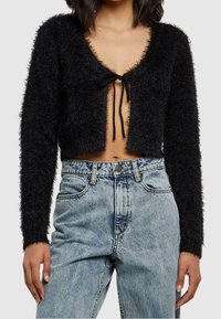 Cardigan noir court en matière douce, avec un décolleté en V profond, des manches longues et une fermeture à nouer à l'avant. Associé à un jean taille haute.