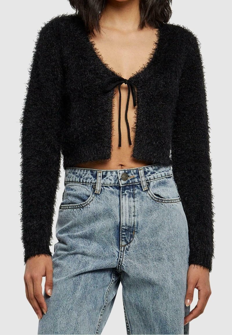 Cardigan noir court en matière douce, avec un décolleté en V profond, des manches longues et une fermeture à nouer à l'avant. Associé à un jean taille haute.