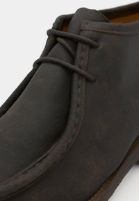 Scarpa chukka in pelle marrone con punta arrotondata, cuciture evidenti e due occhielli per lacci. La superficie ha una finitura leggermente texture.