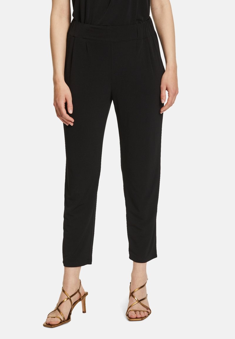 Pantalons noirs fuselés avec une taille élastique, présentant une texture de tissu lisse et des plis à l'avant, associés à des talons métalliques à lanières.