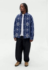 Άνδρας με πλεξούδες μέχρι τους ώμους, φορώντας ένα μπλε πουκάμισο με σχέδια, πάνω από ένα λευκό μπλουζάκι, μαύρα φαρδιά παντελόνια και καφέ παπούτσια, standing against a white background.