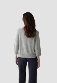 Helles graues Sweatshirt mit Dreiviertelärmeln und geripptem Saum, das ein strukturiertes Strickmuster aufweist. Über dunklen Hosen getragen, Ansicht von hinten.