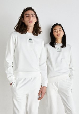 Weiße Sweatshirts mit Rundhalsausschnitt und passenden Hosen, mit einem grünen "LACOSTE"-Logo und Krokodil-Emblem. Weicher Stoff mit gerippten Bündchen.