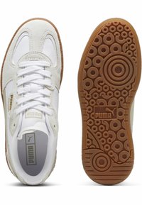 Sneakers blanches en matériaux suède et cuir, avec un design à lacets, semelle extérieure en caoutchouc brun comportant des motifs circulaires, et branding Puma doré.