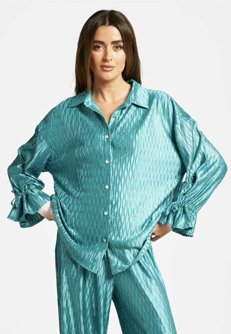 Camisa de seda turquesa con un patrón texturizado de diamantes, que cuenta con cuello y cierre de botones. Corte holgado con detalles de mangas fruncidas.