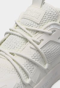 Κοντινό πλάνο άσπρου αθλητικού παπουτσιού από δίχτυ με σταυρωτά κορδόνια και την επωνυμία "Jeans Couture" στη γλώσσα και στη σόλα.