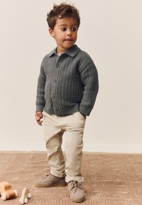 Gray knitted cardigan with a collar, beige pants, and brown shoes. Standing on a textured rug, wooden toy beside. Short, curly hair.
