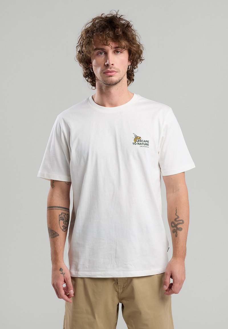 Valkoinen puuvillapaita, jossa on pieni graafinen kuva mehiläisestä ja teksti "PAETAA LUONTOON KALIFORNIA" vasemmassa rintakehässä. Pyöreä kaula-aukkokdesign.