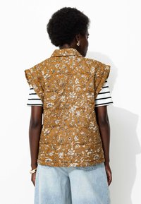 Personne portant un gilet sans manches matelassé à fleurs moutarde par-dessus une chemise à rayures à manches courtes et un jean taille large bleu clair, de dos.
