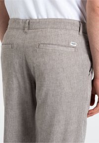 Jasnobeżowe lniane spodnie o gładkiej fakturze, wyposażone w dwie tylne kieszenie oraz metkę po prawej stronie. Widoczny regulowany pas w talii.